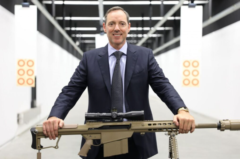 NIOA CEO Rob Nioa standing in front of a sniper rifle, wearing a blue suit and smiling at the camera in war criminal glee.